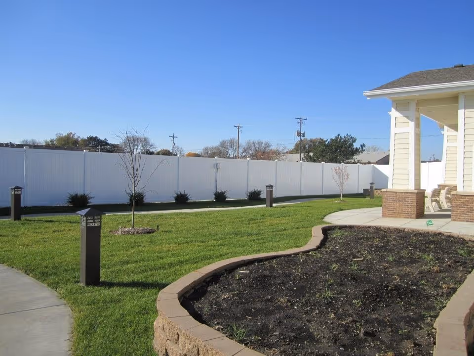 Outdoor area of Hansen House featuring a well-maintained lawn, a curved garden bed with soil, a paved walkway, small trees, and a white privacy fence under a clear blue sky. Part of a building with a covered porch and chairs is visible on the right.