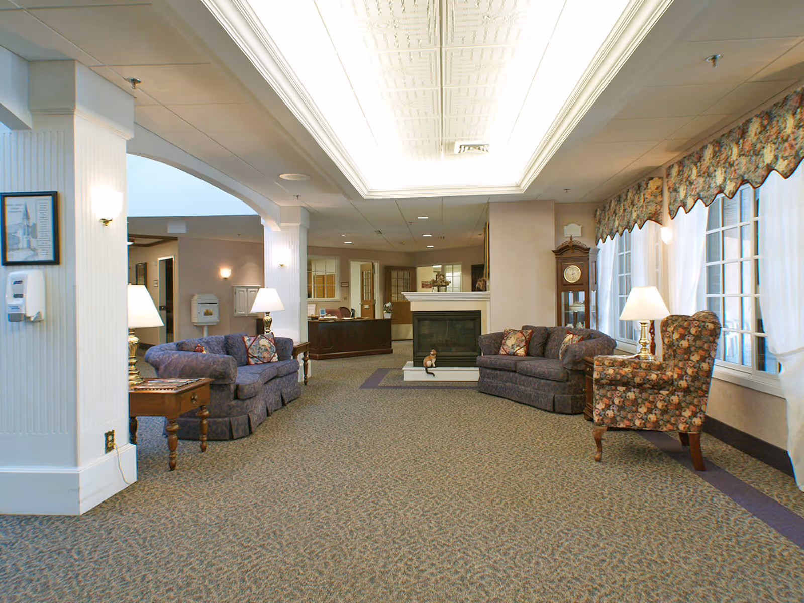 Spacious senior living lounge with sofas and armchairs arranged around a central fireplace, lamps, and large windows with valances.