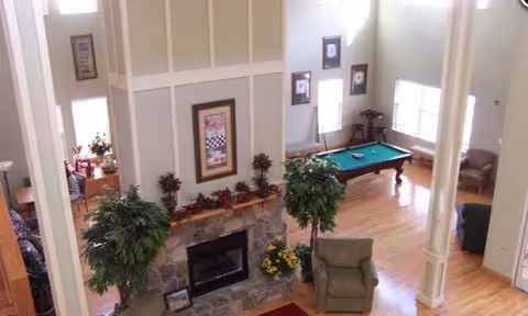 Spacious living area with high ceilings featuring a stone fireplace decorated with plants and flowers. There are two large potted plants near the fireplace, a green armchair, a pool table with cues, and several windows letting in natural light. Additional seating and a dining area are visible in the background.