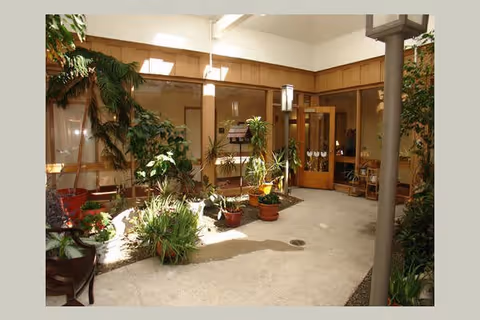 Indoor courtyard area with various potted plants and greenery, surrounded by wooden framed windows and doors leading to interior rooms. The space is well-lit with natural light and has a clean, open floor with some gravel along the edges.