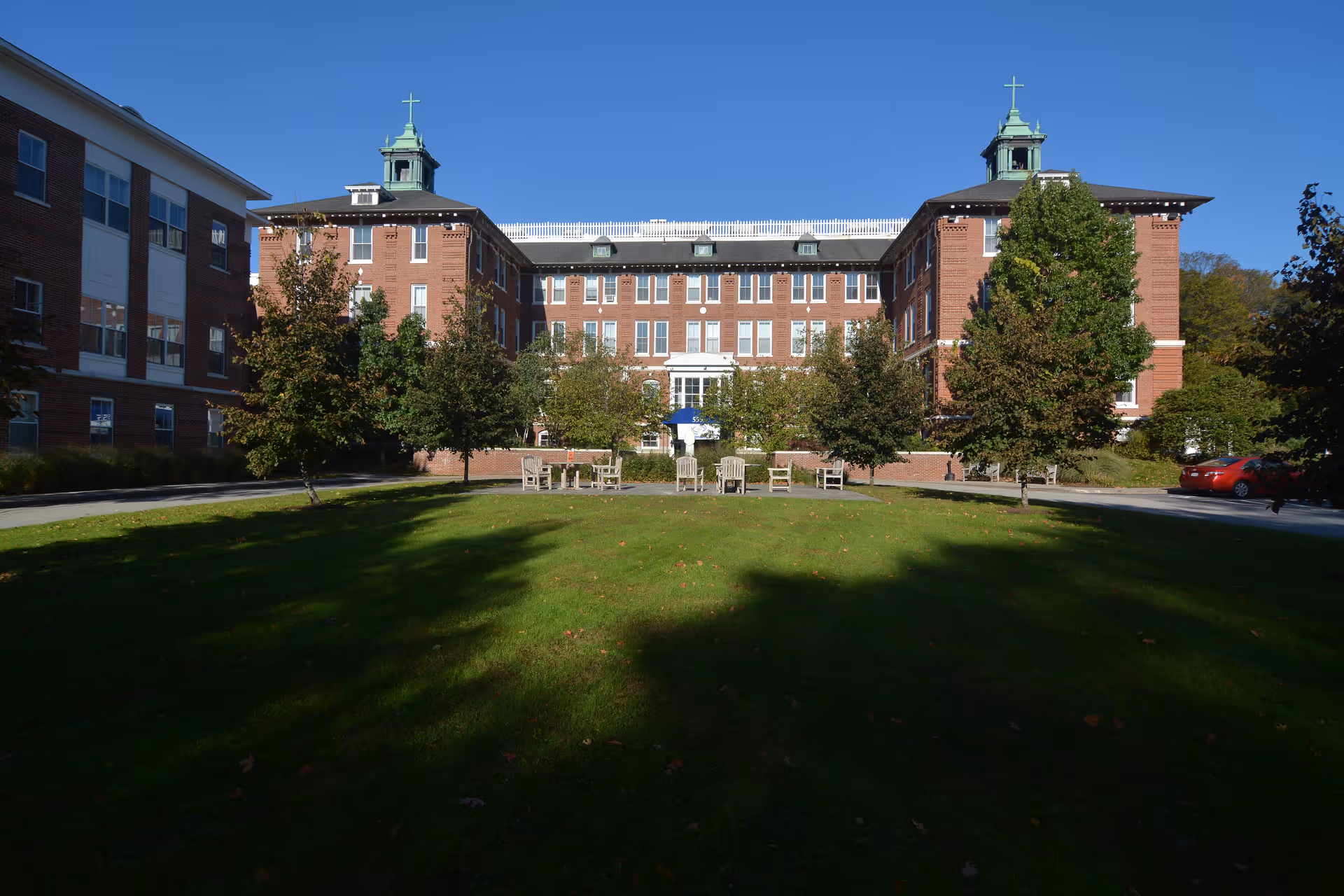 Front exterior view of a large brick building with multiple windows and two cupolas on the roof, surrounded by trees and a green lawn with outdoor seating under a clear blue sky.