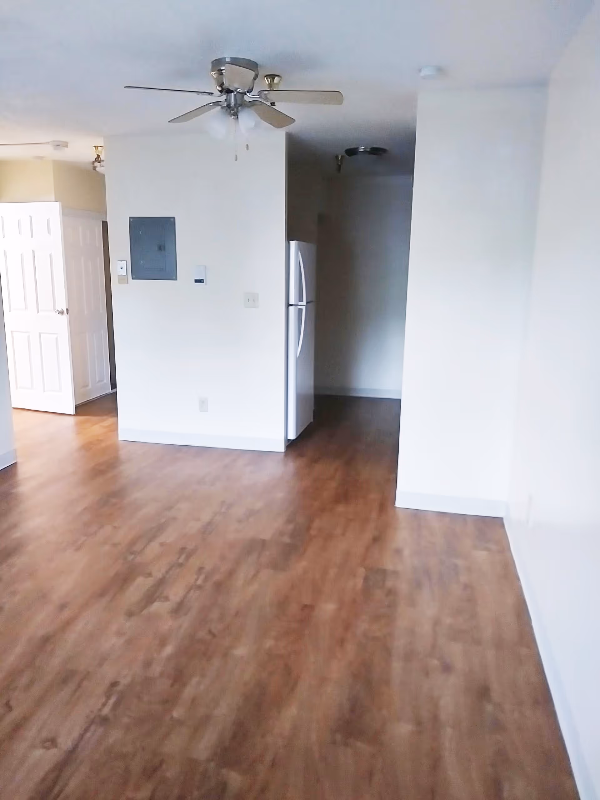 Empty room with wood flooring, white walls, a ceiling fan with lights, and a white refrigerator partially visible in a small kitchen area. There is an open door leading to another room on the left side.