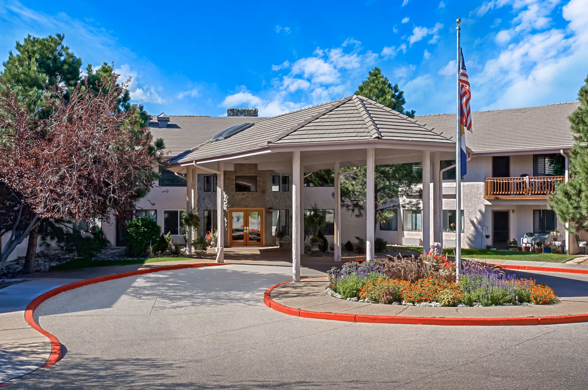 Exterior view of Solista Colorado Springs senior living facility showing the entrance with a covered driveway, a circular flower bed with colorful flowers, an American flag on a flagpole, and surrounding trees under a blue sky with some clouds.