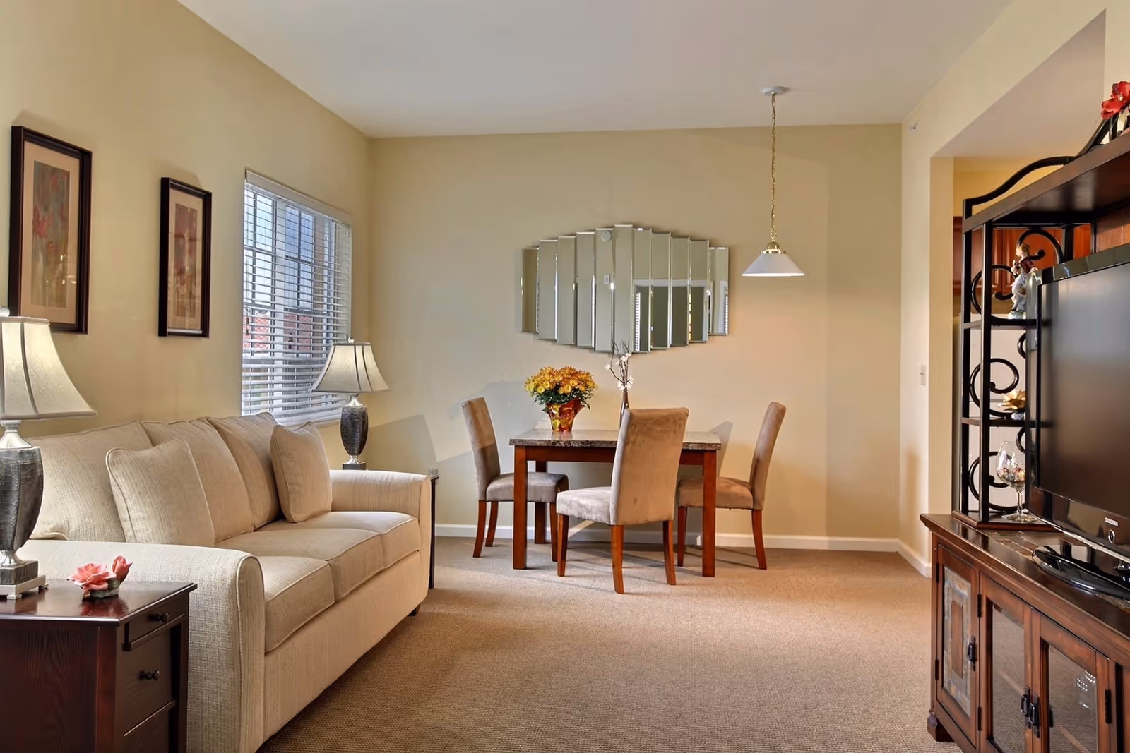 A cozy living and dining area with a beige sofa, two table lamps, a wooden side table, a dining table with four upholstered chairs, a decorative mirror on the wall, and a wooden entertainment center with a TV and decorative items.