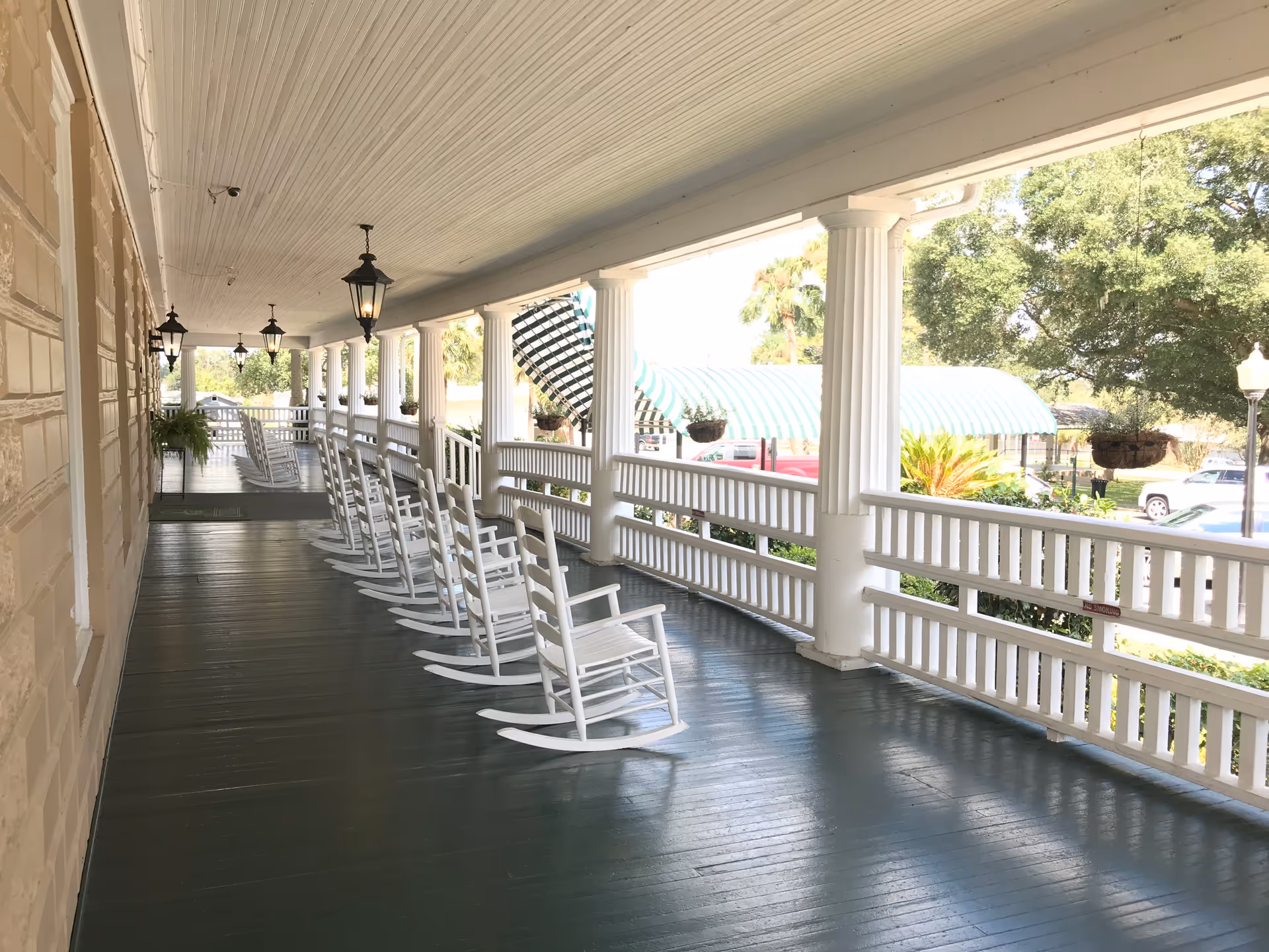 A long covered porch with white wooden rocking chairs lined up along the railing. The porch has white columns and hanging lantern-style lights. Outside, there are trees, parked cars, and a green and white striped awning.