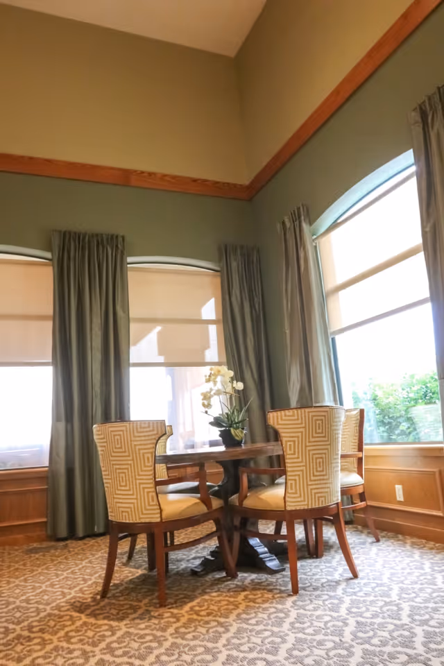 Small dining area with a round wooden table, four patterned chairs, tall windows with curtains, and a floral centerpiece.