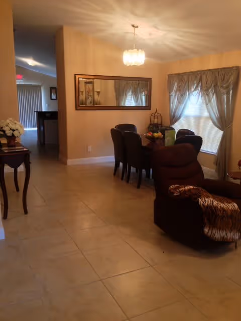 Open-plan interior showing a dining table with chairs under a chandelier, a recliner in the foreground, and a window with drapes.