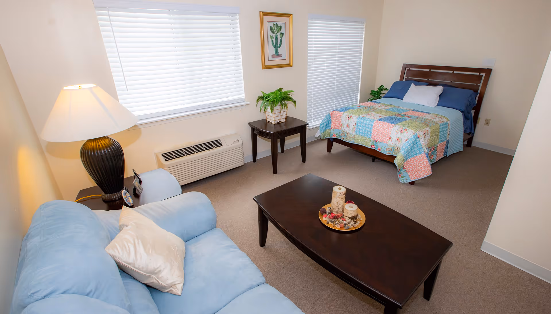 A cozy room with a light blue sofa with a white pillow, a dark wooden coffee table with decorative candles, a side table with a lamp and a plant, and a bed with a colorful patchwork quilt. The room has two large windows with white blinds and light beige walls.