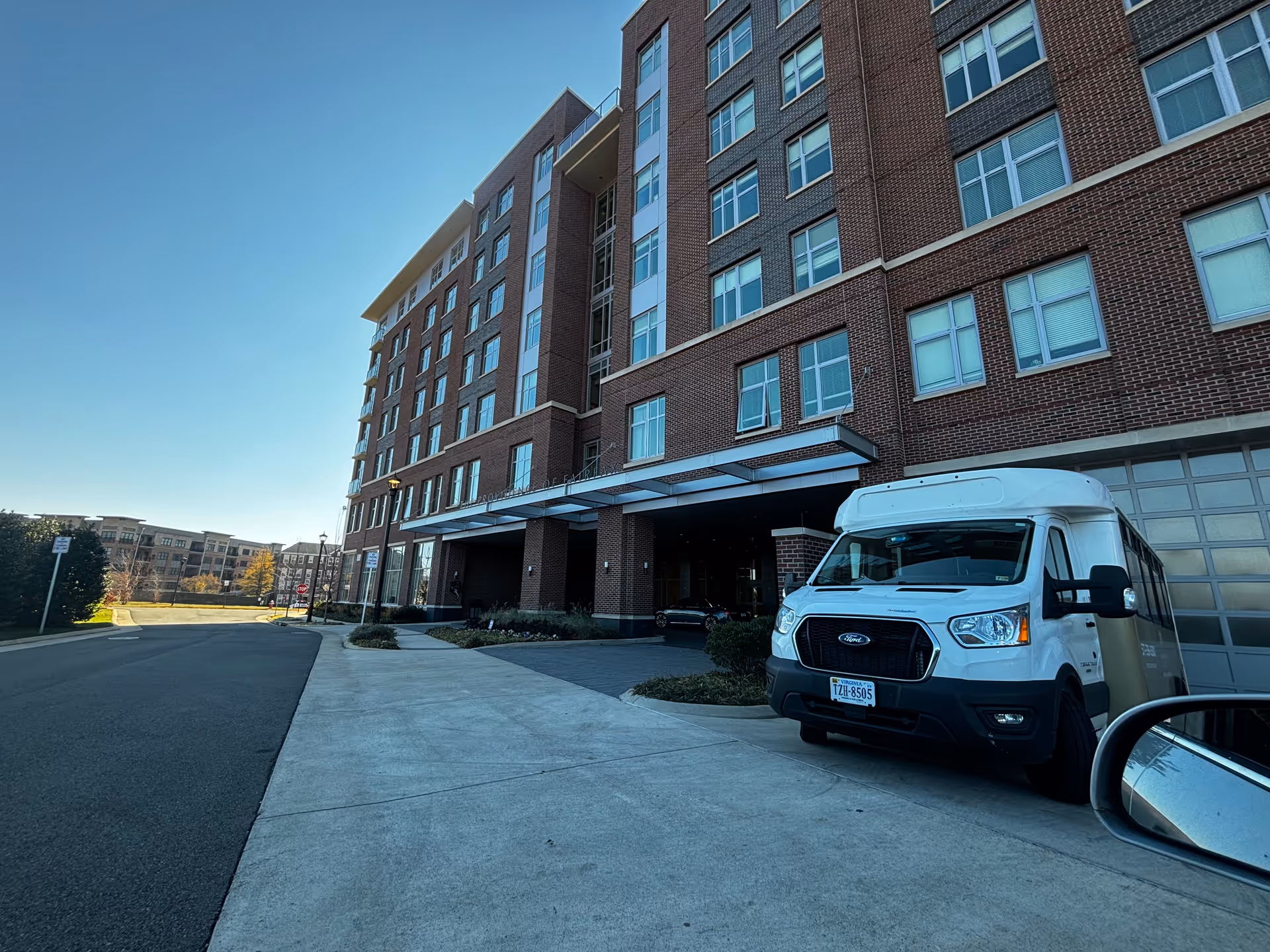 Exterior view of The Providence Fairfax building showing a multi-story brick facade with numerous windows. A white shuttle van is parked on the driveway near the entrance. The sky is clear and blue.