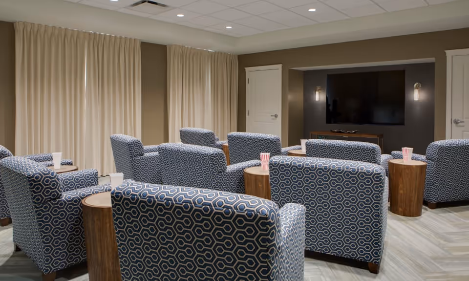 A cozy living room area with multiple blue patterned armchairs arranged in rows facing a large flat-screen TV mounted on a dark accent wall. Small wooden side tables are placed between the chairs, each holding a striped popcorn container. The room has beige walls, light-colored curtains covering the windows, and a light wood floor.