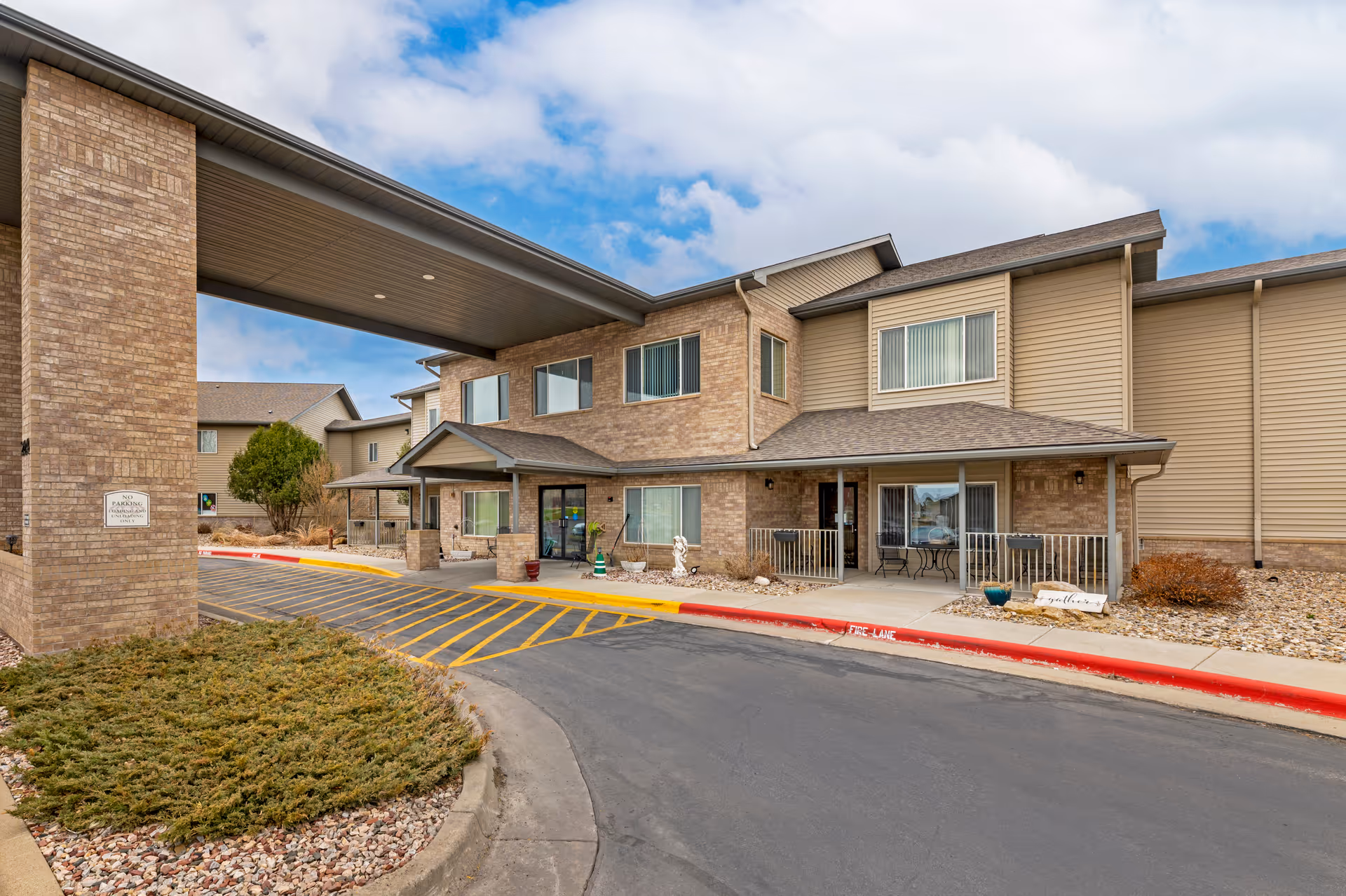 Exterior view of Arrowhead Lodge Senior Living facility showing a two-story building with brick and siding facade, a covered entrance with a driveway, and some landscaping with bushes and rocks under a partly cloudy sky.