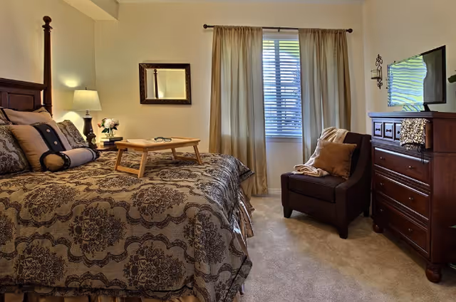 A cozy bedroom with a large bed covered in a patterned bedspread, a wooden tray with glasses on the bed, a bedside table with a lamp and flowers, a mirror on the wall, a window with curtains, a cushioned armchair with pillows, and a wooden dresser with a TV on top.