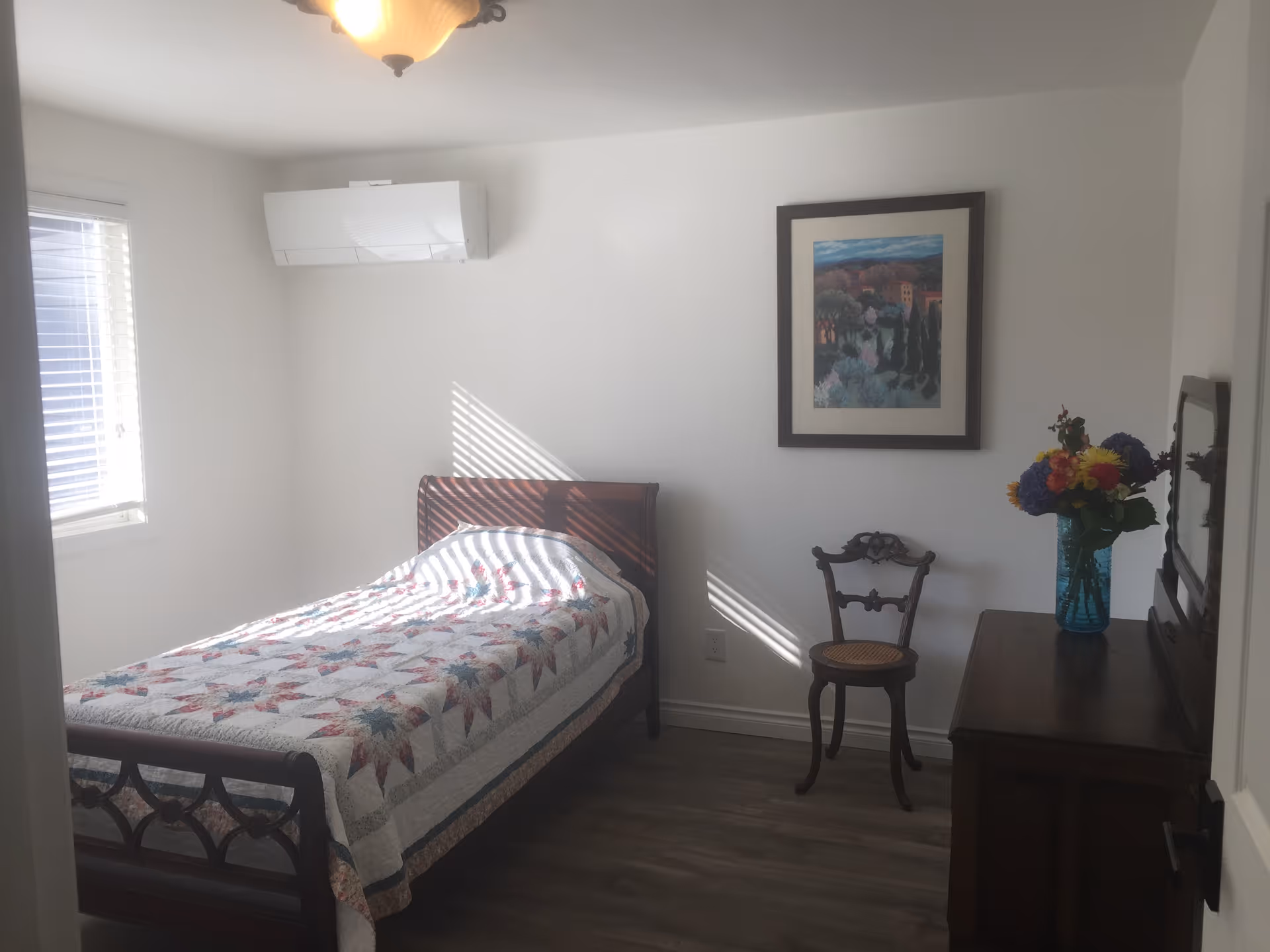 A small, bright bedroom with a single bed covered with a colorful quilt. There is a wooden chair with a woven seat next to a wooden dresser that has a vase with colorful flowers on it. A framed painting hangs on the white wall above the chair. A window with blinds lets in natural light, and an air conditioning unit is mounted on the wall above the bed.