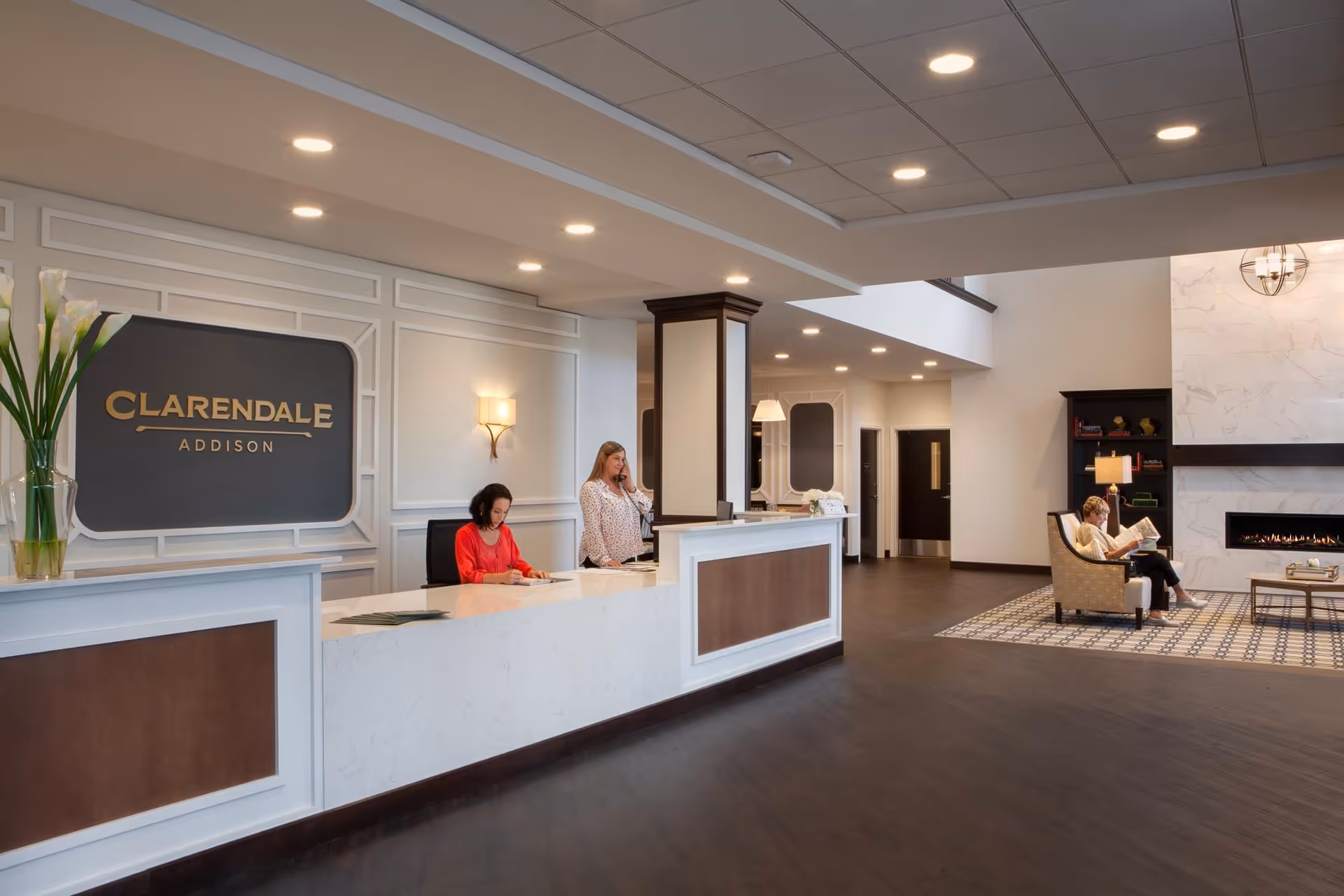 Reception area of Clarendale of Addison with two receptionists behind a white and brown front desk. A woman is sitting in a chair reading a book near a modern fireplace in a cozy seating area. The space is well-lit with recessed ceiling lights and wall sconces, featuring a clean and elegant design.