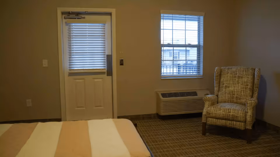 Interior view of a room with a bed partially visible in the foreground, a door with closed blinds, a window with blinds showing an outside view, a wall-mounted air conditioning unit below the window, and a patterned armchair placed near the corner.