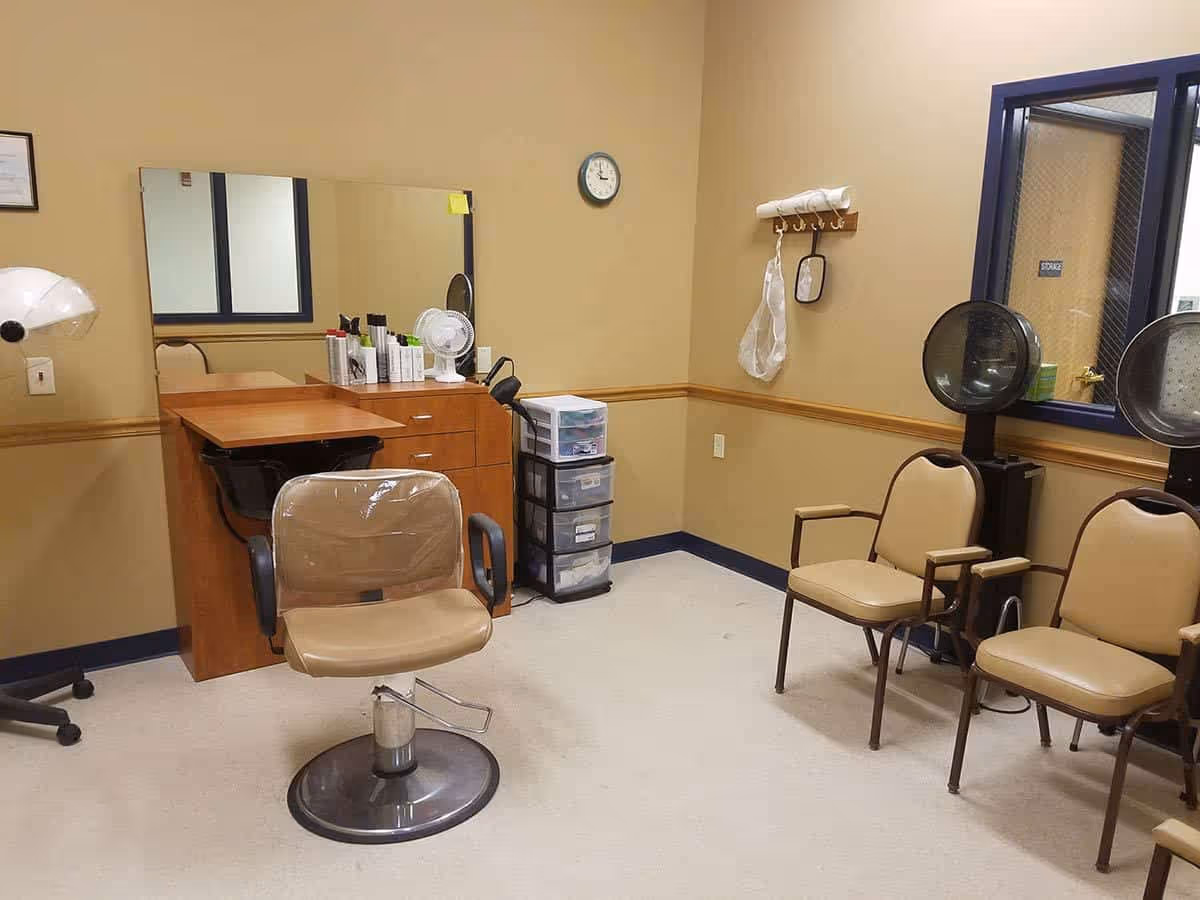 Interior of a hair salon area in a senior living facility with a salon chair in front of a wooden counter and mirror. There are hair care products and a small fan on the counter. On the right side, there are two beige chairs with armrests and two hair dryers. The walls are beige with a wooden chair rail and a clock above the counter.