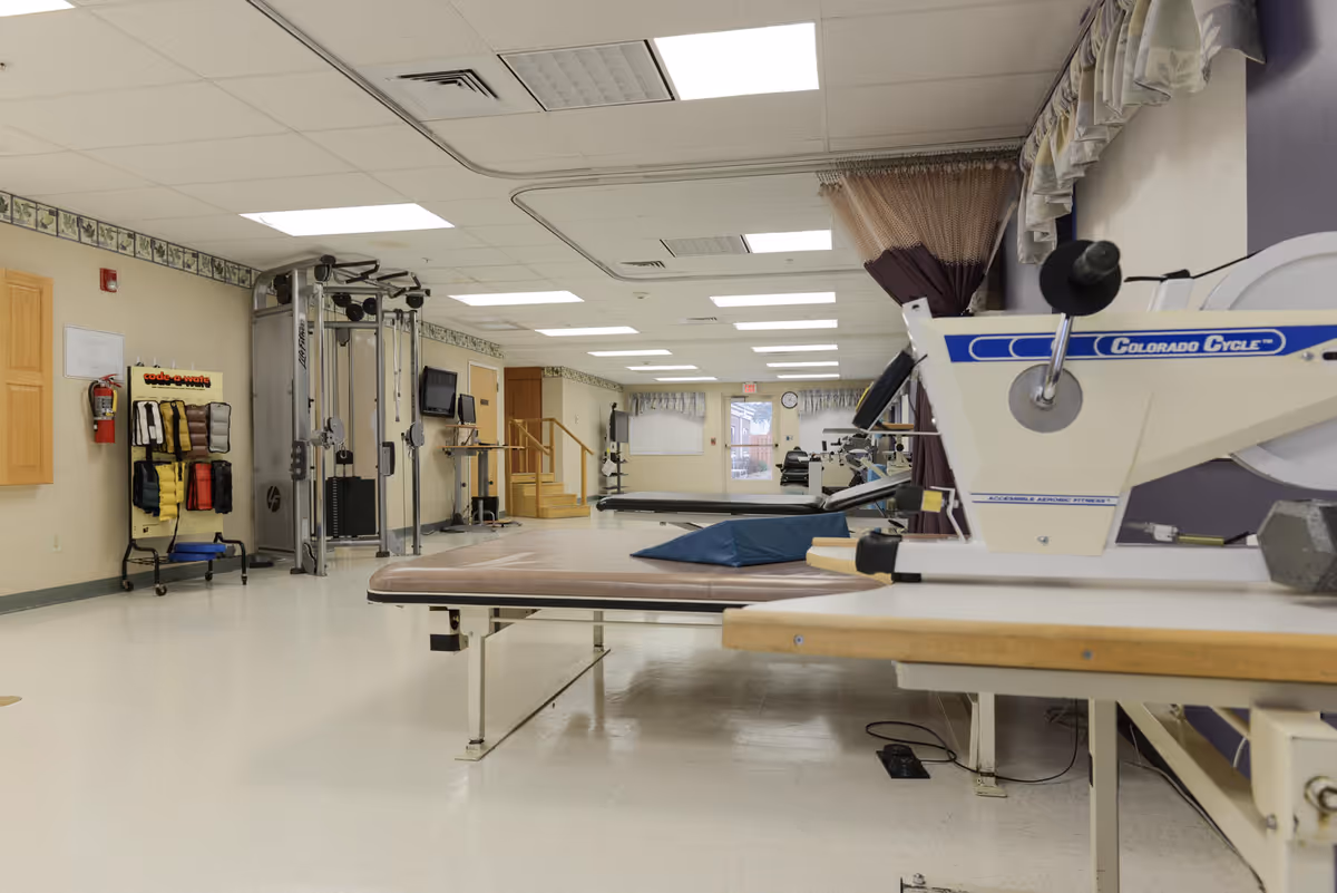 Interior view of a rehabilitation and nursing facility room with exercise equipment including a Colorado Cycle machine, weight racks, therapy tables, and a multi-functional exercise machine. The room has a clean, bright appearance with white floors, beige walls, and fluorescent ceiling lights.