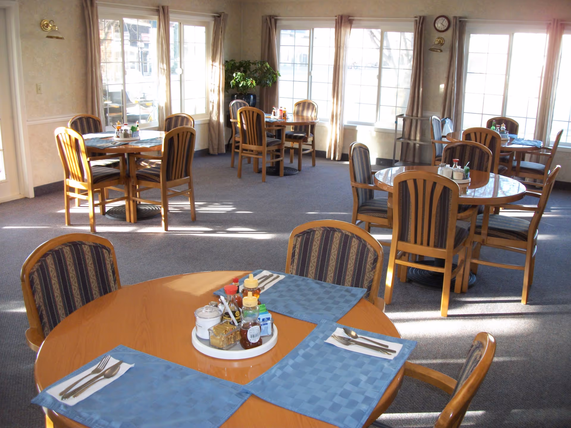 Sunlit dining room with several round wooden tables and chairs set with placemats and condiments.