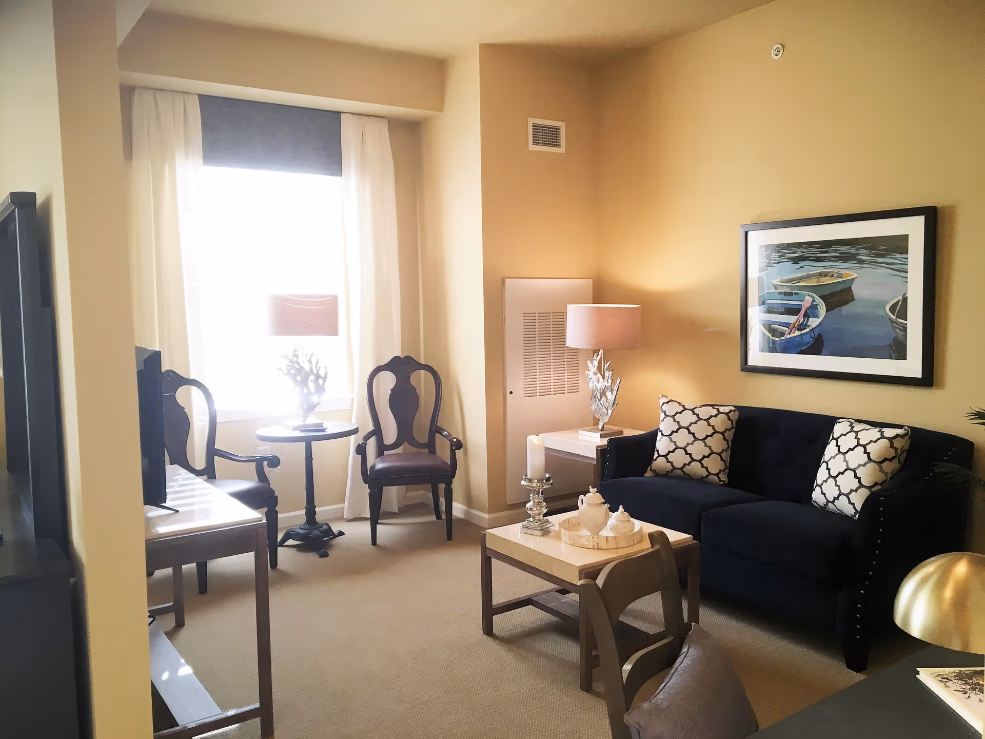A cozy living room with a dark blue sofa adorned with two patterned cushions, a coffee table with decorative items, two wooden chairs with dark cushions near a window with white curtains, a side table with a lamp, and a framed picture of boats on the water hanging on the wall.