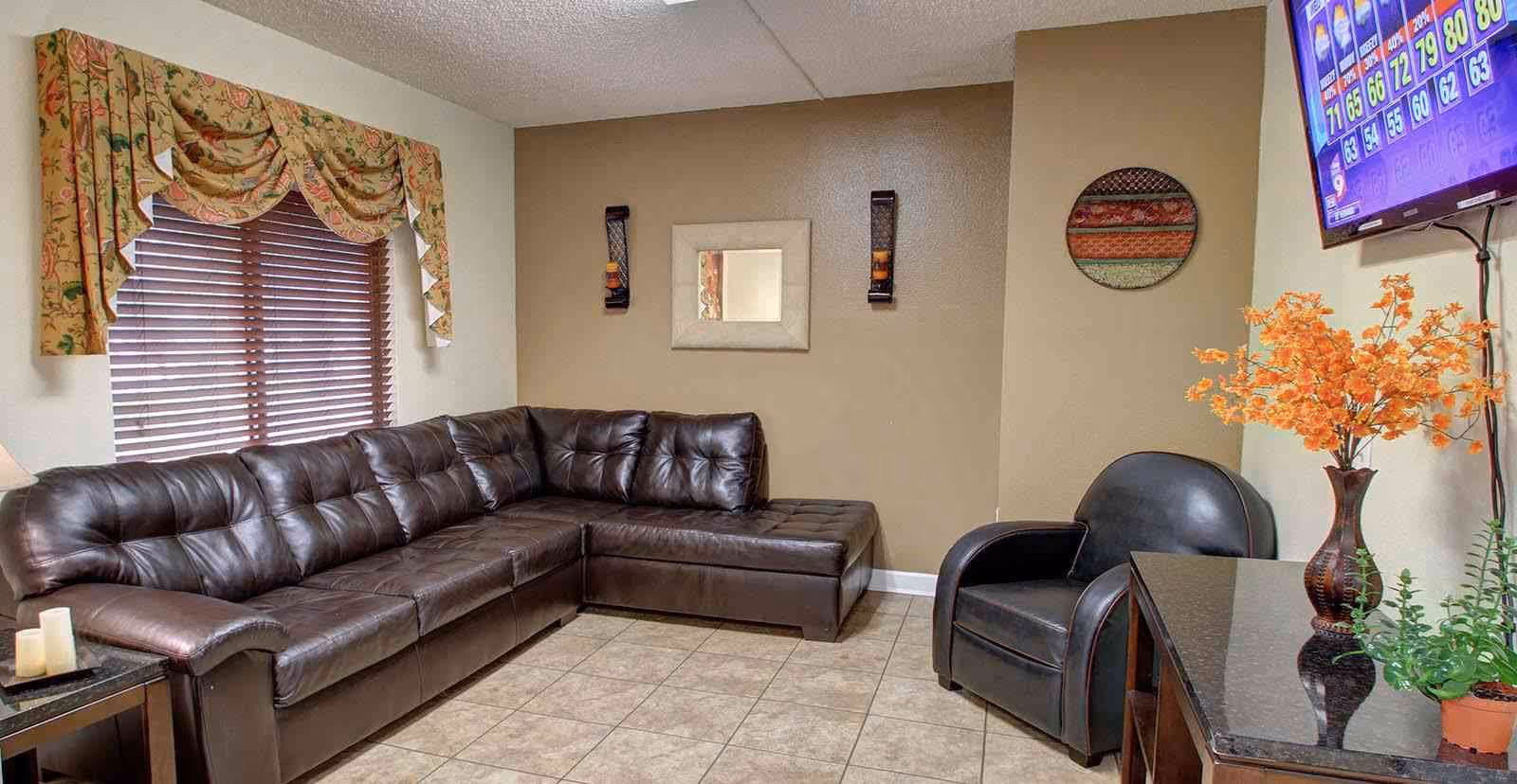 A cozy living room with a large dark brown leather sectional sofa, a black leather armchair, and a black granite-top table with a vase of orange flowers and a small green plant. The walls are painted beige and white, with a small square mirror and two candle sconces on the wall. A window with wooden blinds and floral valance is on the left side, and a flat-screen TV is mounted on the right wall displaying a colorful bingo game screen.