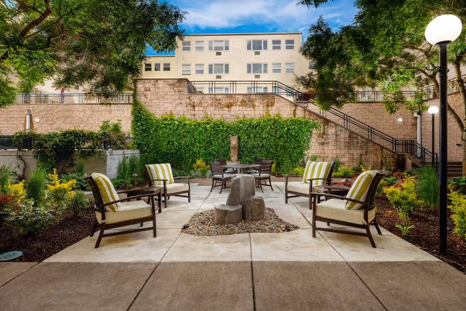 Outdoor seating area with cushioned chairs arranged around a small stone fire pit, surrounded by landscaped greenery and trees, with a multi-story building and stairs in the background under a blue sky.