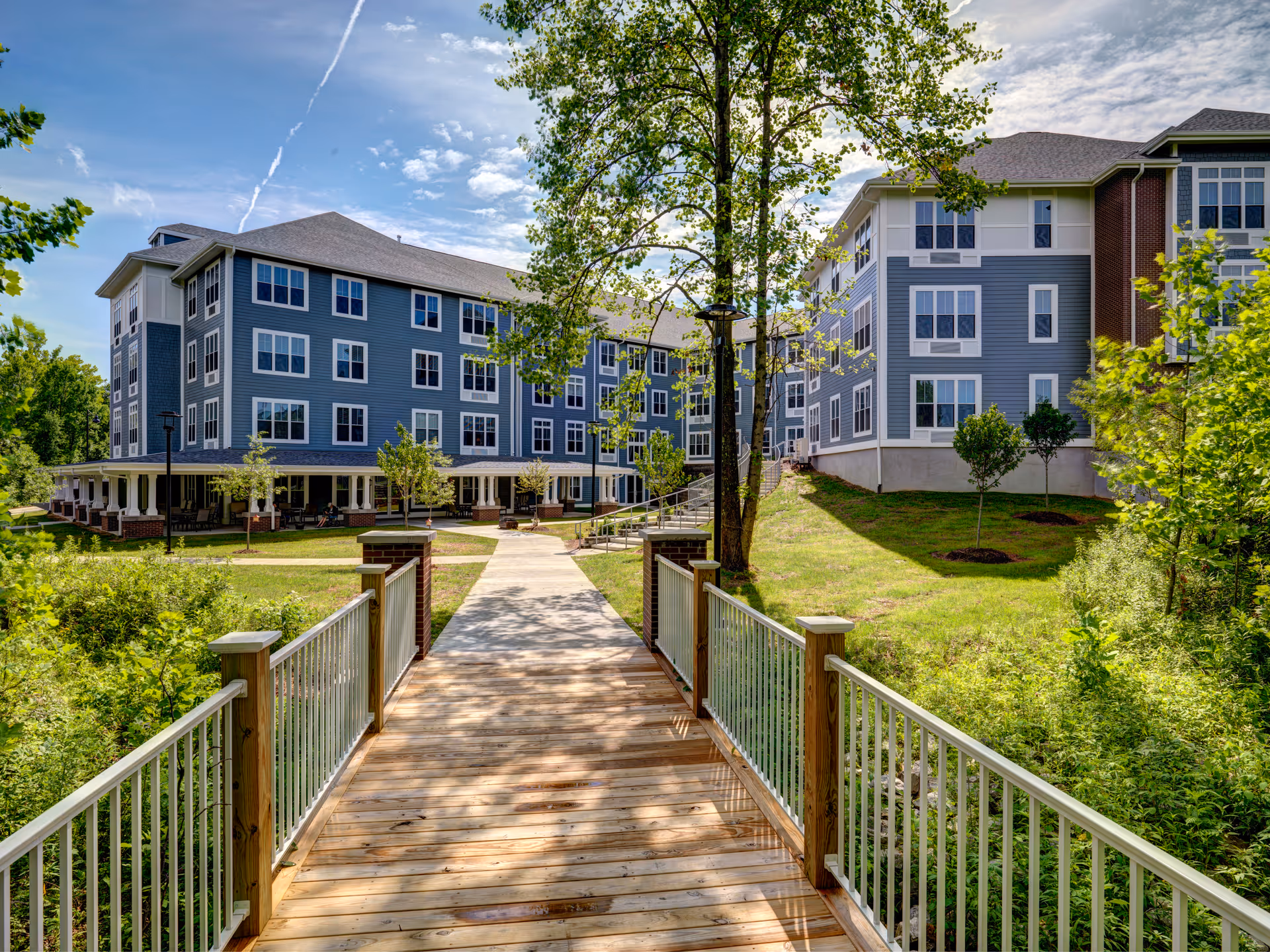 A wooden pedestrian bridge with white railings leads to a paved walkway surrounded by greenery and trees, approaching a large multi-story residential building with blue and white siding and numerous windows under a partly cloudy sky.