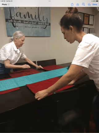 Two women setting a dining table with red placemats and a turquoise table runner in a room with a wall decoration that reads 'family side by side - hand in hand - heart to heart'.