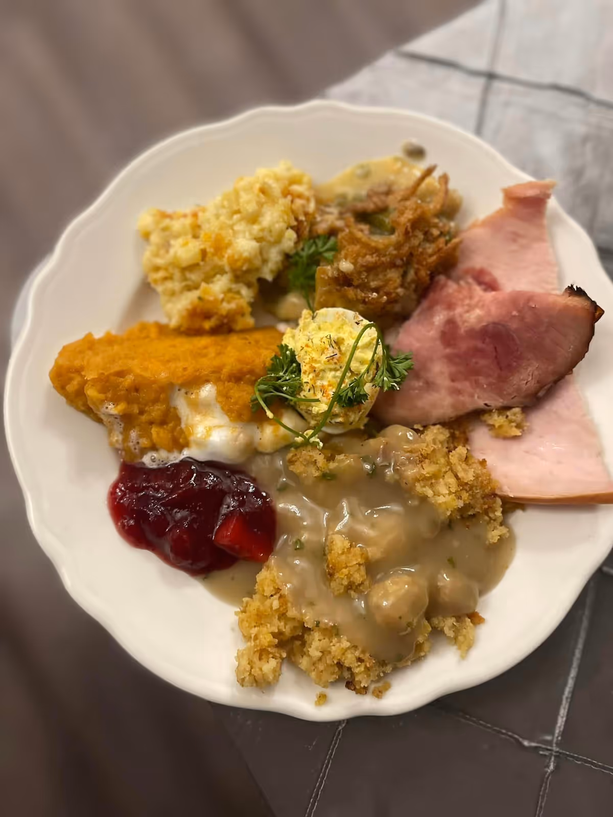 A white plate filled with a variety of food including slices of ham, stuffing with gravy, cranberry sauce, a deviled egg garnished with parsley, a casserole dish, and a breaded item with melted cheese.