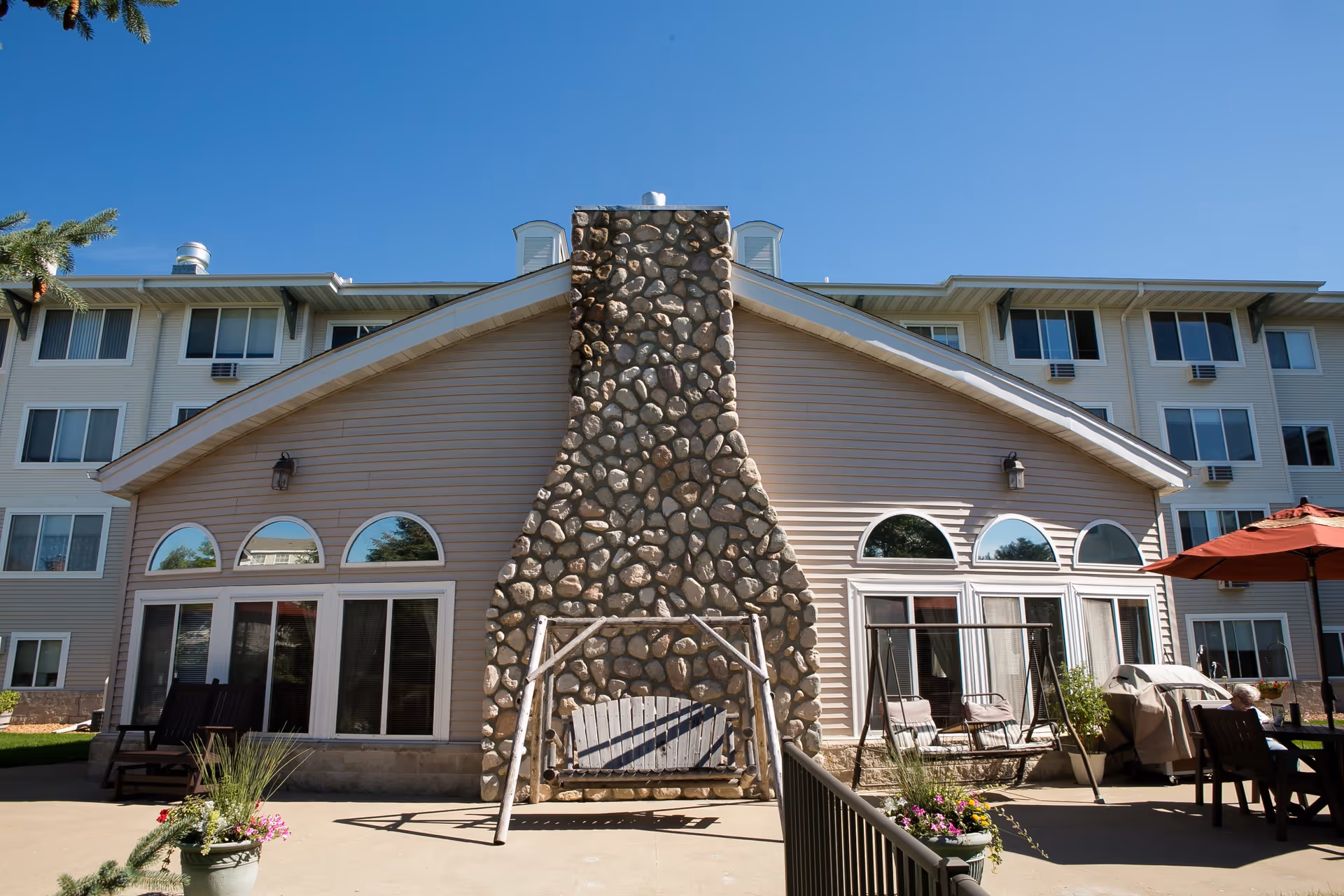 Outdoor patio area of Westridge Retirement Community featuring a large stone fireplace with a wooden swing attached. The building has beige siding with arched windows and multiple floors. There are patio chairs, a table with a red umbrella, potted plants, and a clear blue sky above.