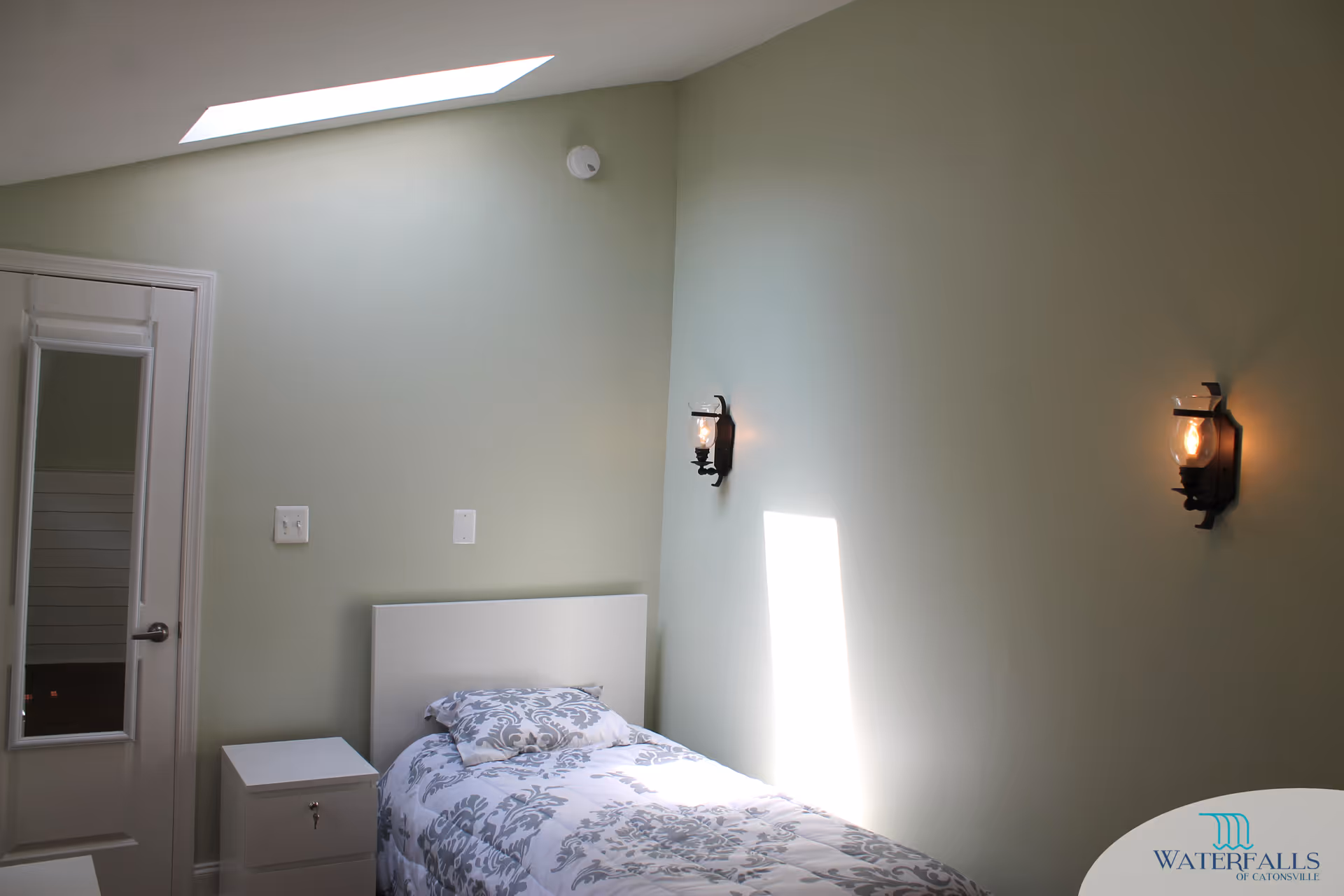 A small bedroom with a single bed featuring a white and gray patterned comforter and pillow. The room has light green walls, two wall-mounted lantern-style lights, a skylight, a white nightstand with a drawer and a door, and a door with a full-length mirror attached.