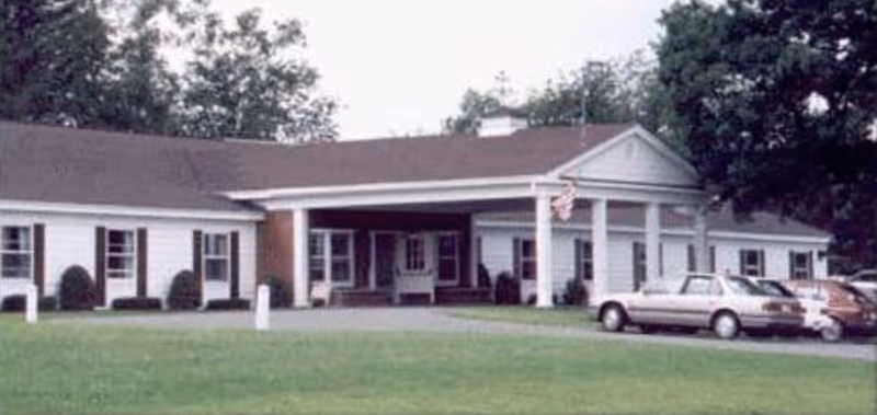 Single-story white nursing home with a covered portico supported by columns, an American flag, a lawn in front, and several parked cars.