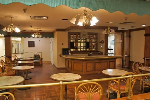 Interior view of a common area in an assisted living facility featuring multiple round tables with chairs, a wooden reception or bar counter in the center, and several ceiling light fixtures with ornate designs. The room has a warm color palette with beige walls and wooden flooring.