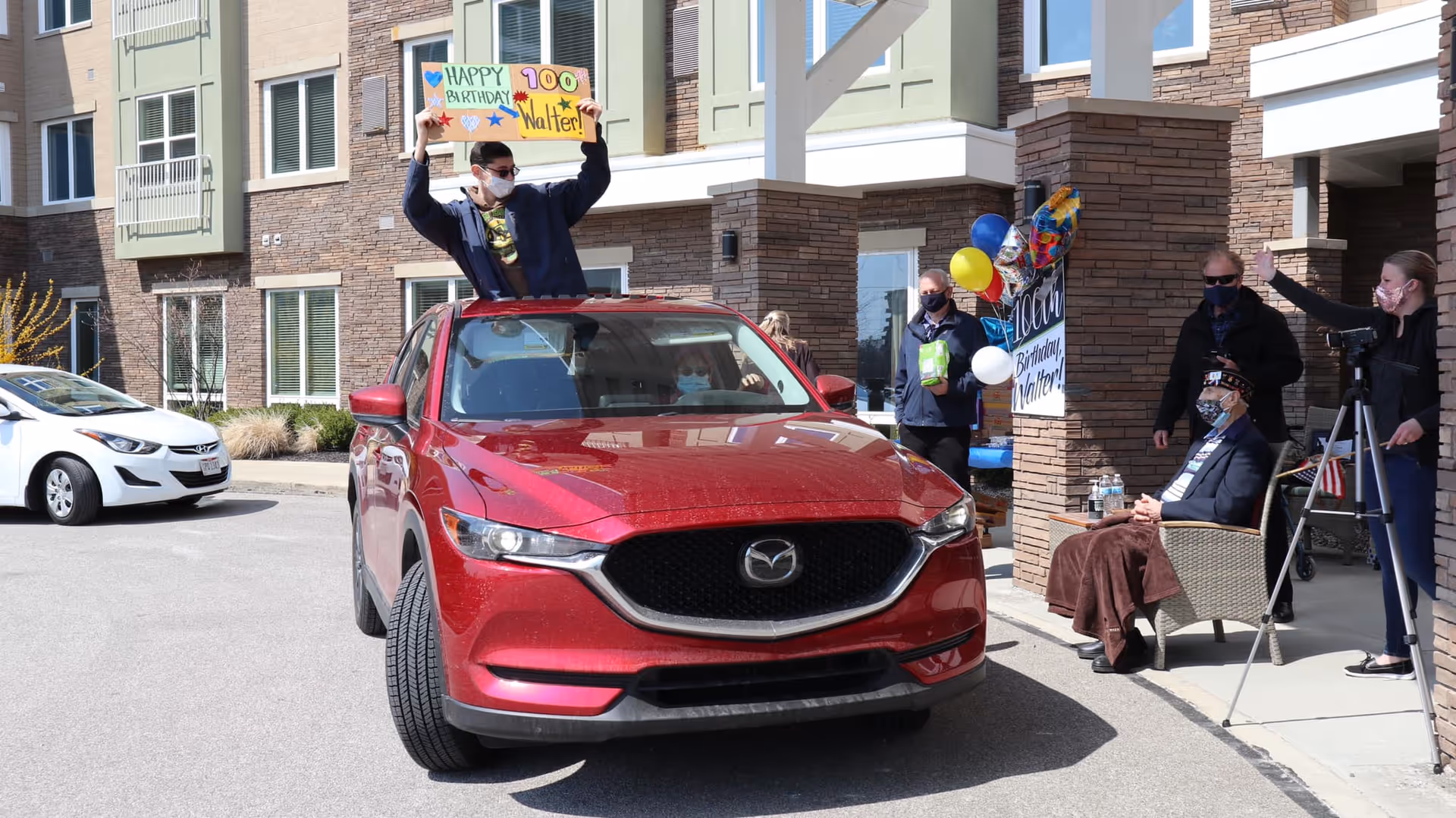 A man wearing a mask is standing through the sunroof of a red Mazda SUV holding a colorful sign that says 'Happy 100th Birthday Walter!' Outside a senior living facility, several people wearing masks stand nearby, including an elderly man sitting in a chair with a blanket over his legs. There are balloons and another birthday sign in the background.