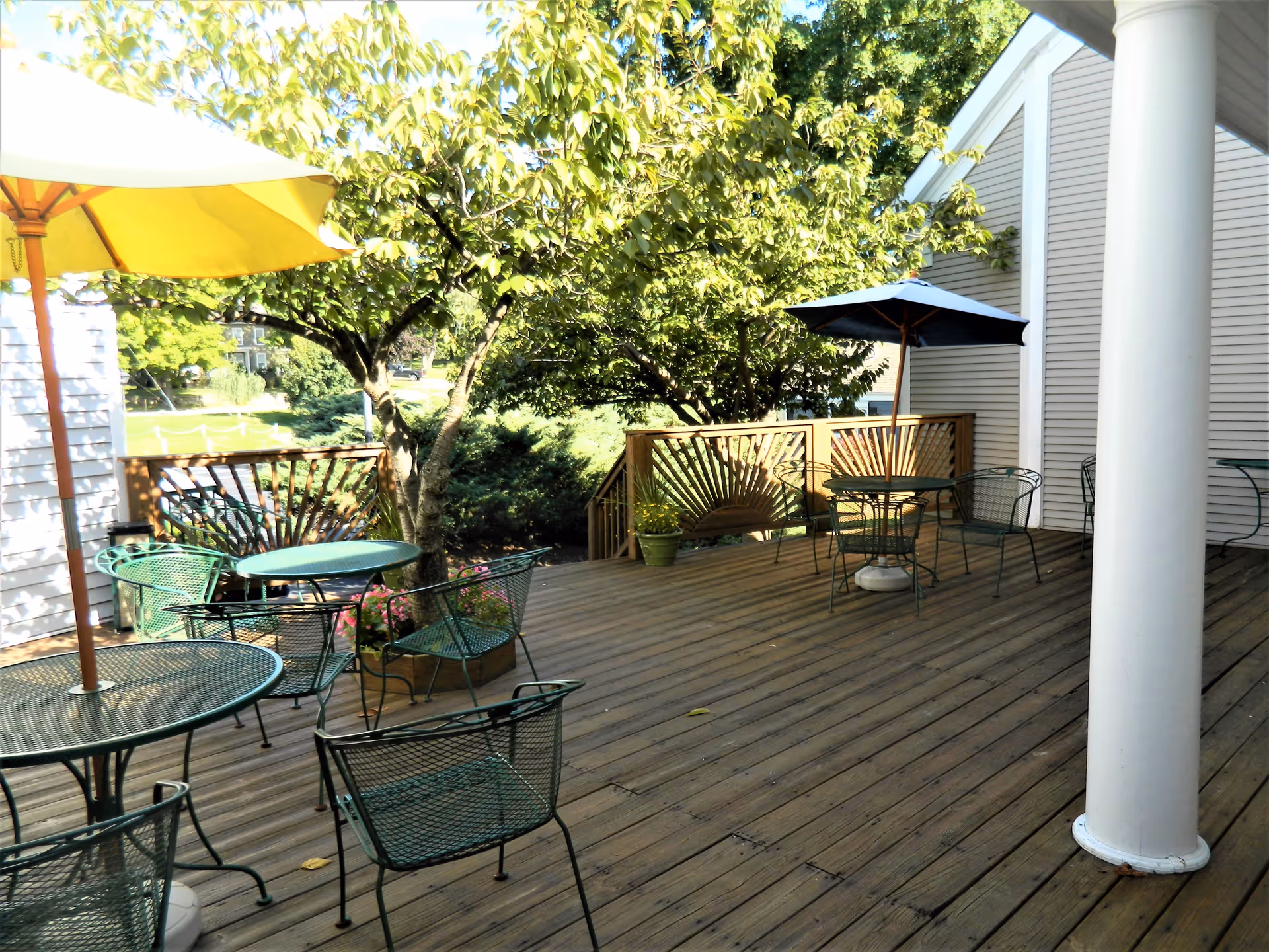 Outdoor wooden deck area with several green metal tables and chairs, two umbrellas providing shade, surrounded by trees and adjacent to a light-colored building with a white column.