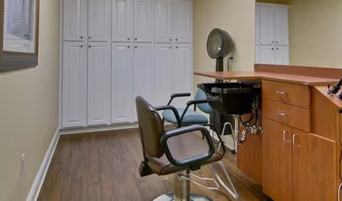 Interior view of a salon area in a senior living facility featuring two salon chairs, a hair dryer, wooden cabinetry with a sink, and white storage cabinets against a beige wall with wood flooring.