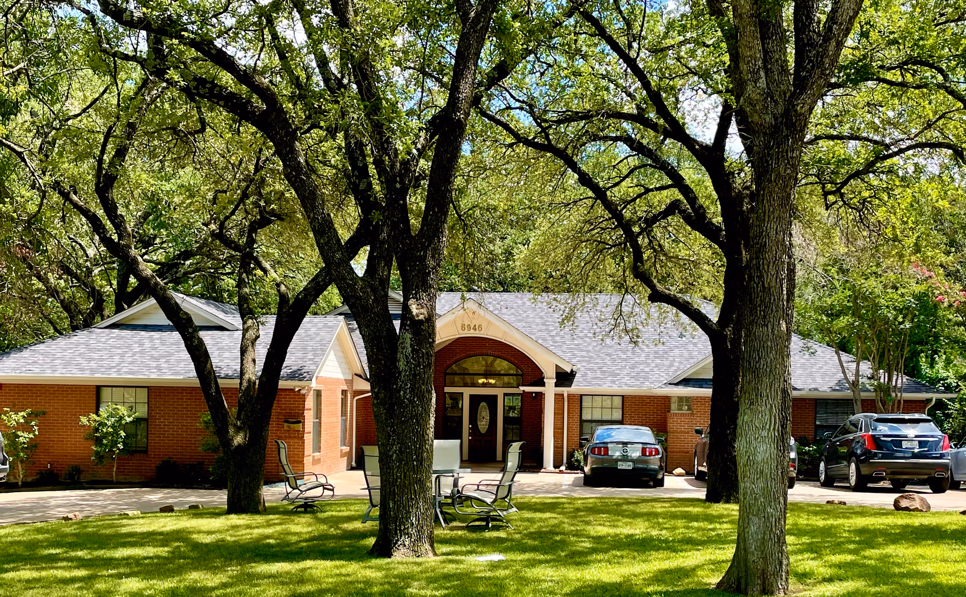 Front exterior view of Magnolia Assisted Living Care Home, a single-story brick building with a gray shingled roof, surrounded by large trees and green lawn. There are several parked cars in the driveway and outdoor chairs on the lawn.