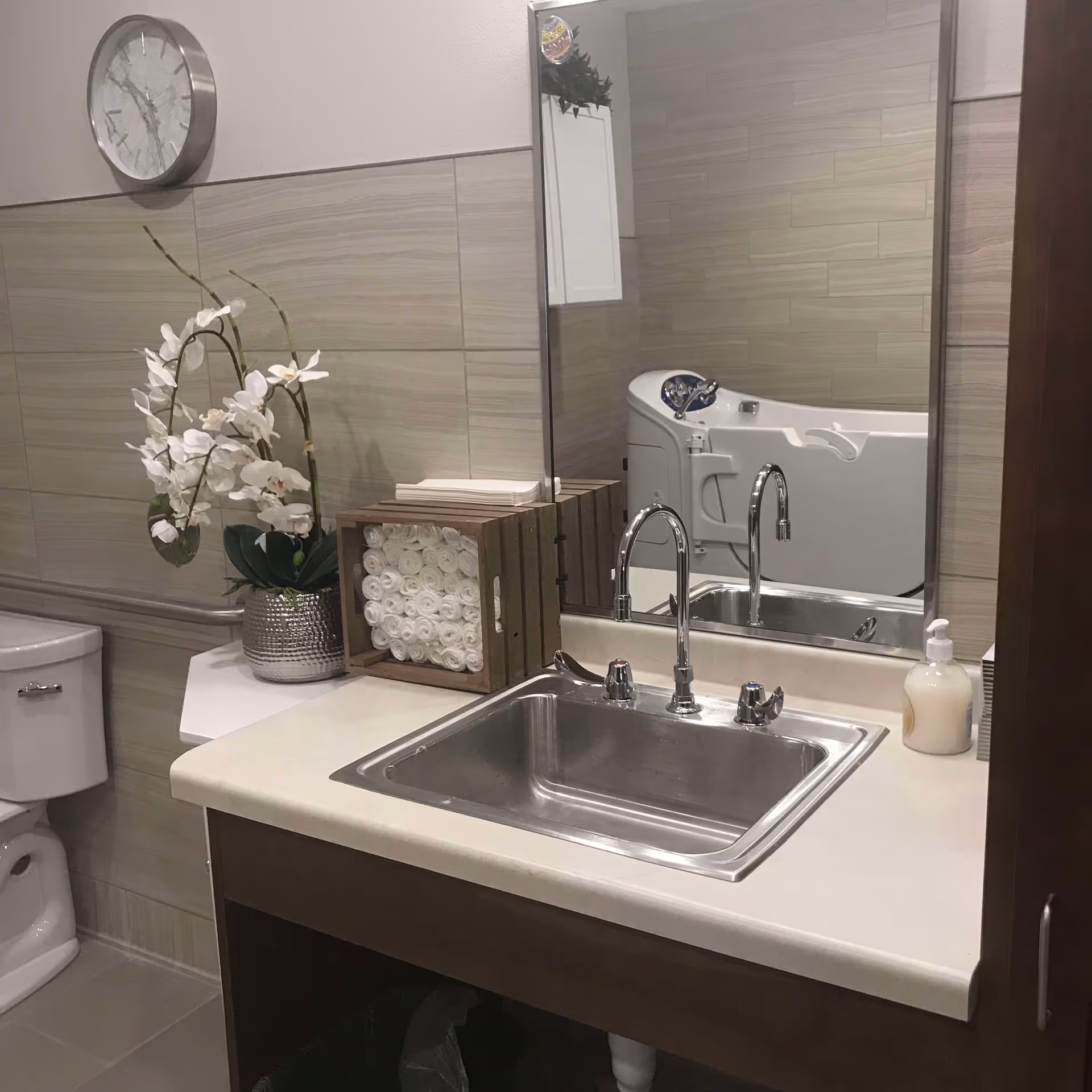 A bathroom vanity with a stainless sink and faucet, mirror, decorative orchids and rolled towels, with a toilet and walk-in tub visible.