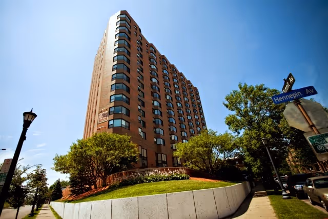 Low-angle view of a multi-story brick apartment building with trees, a grassy raised bed, and a sidewalk under a clear blue sky.