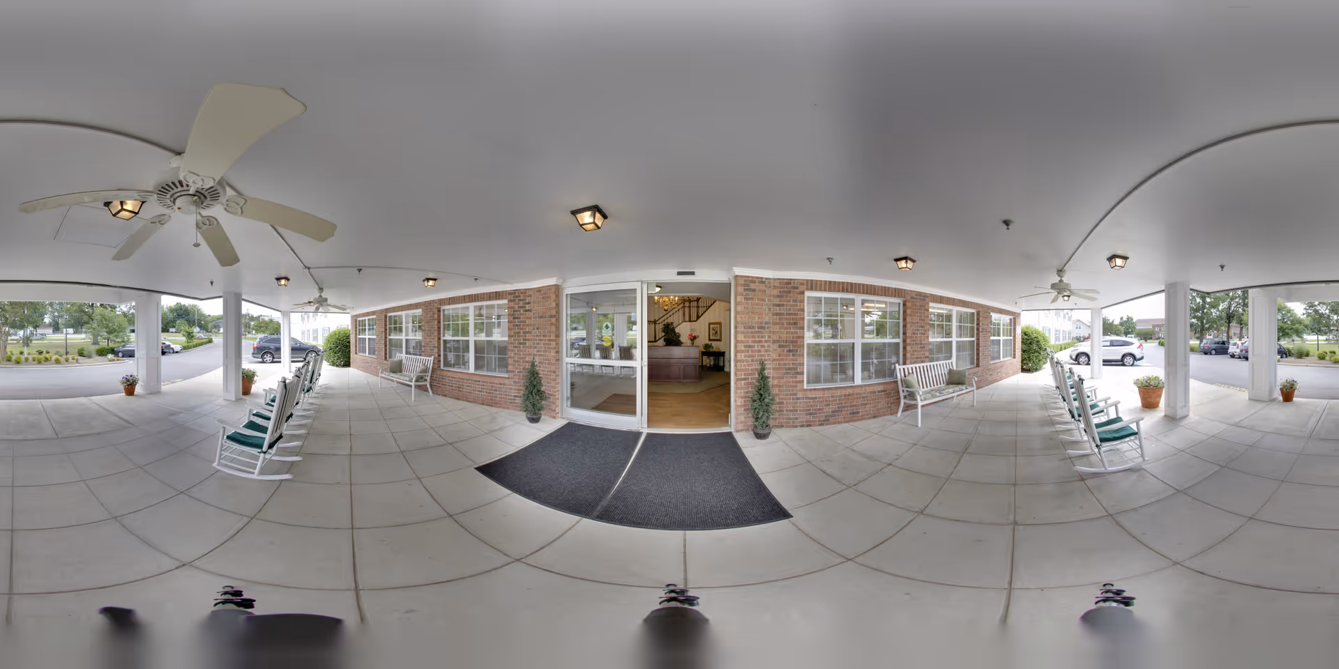 Covered entrance area of a senior living facility with rocking chairs and benches along the brick walls, ceiling fans overhead, potted plants, and a glass door leading inside to a reception area.