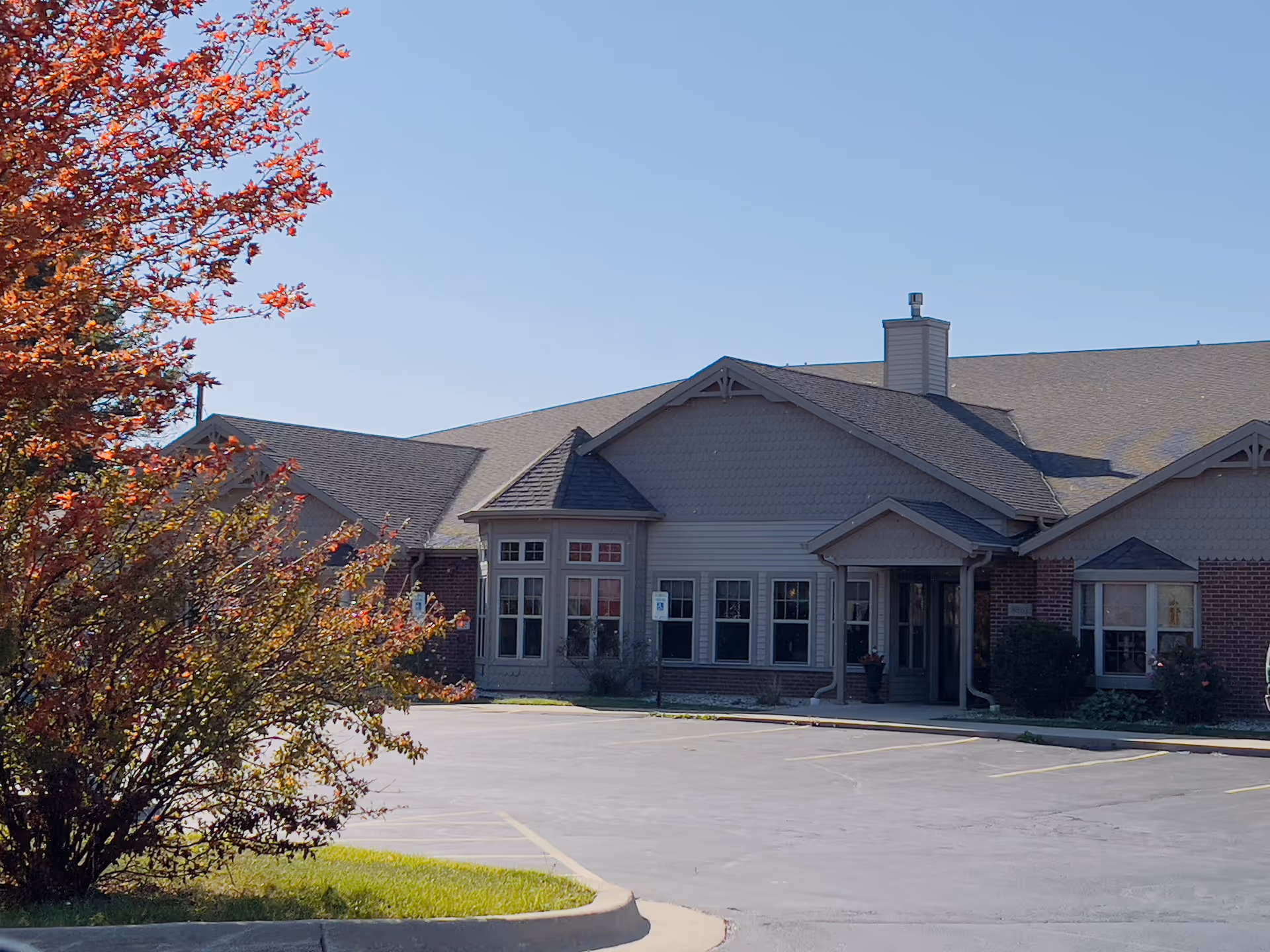 Exterior view of a single-story senior living facility building with a brick and siding facade, multiple windows, and a small covered entrance. There is a parking lot in front with marked parking spaces and a tree with red and orange leaves on the left side of the image under a clear blue sky.