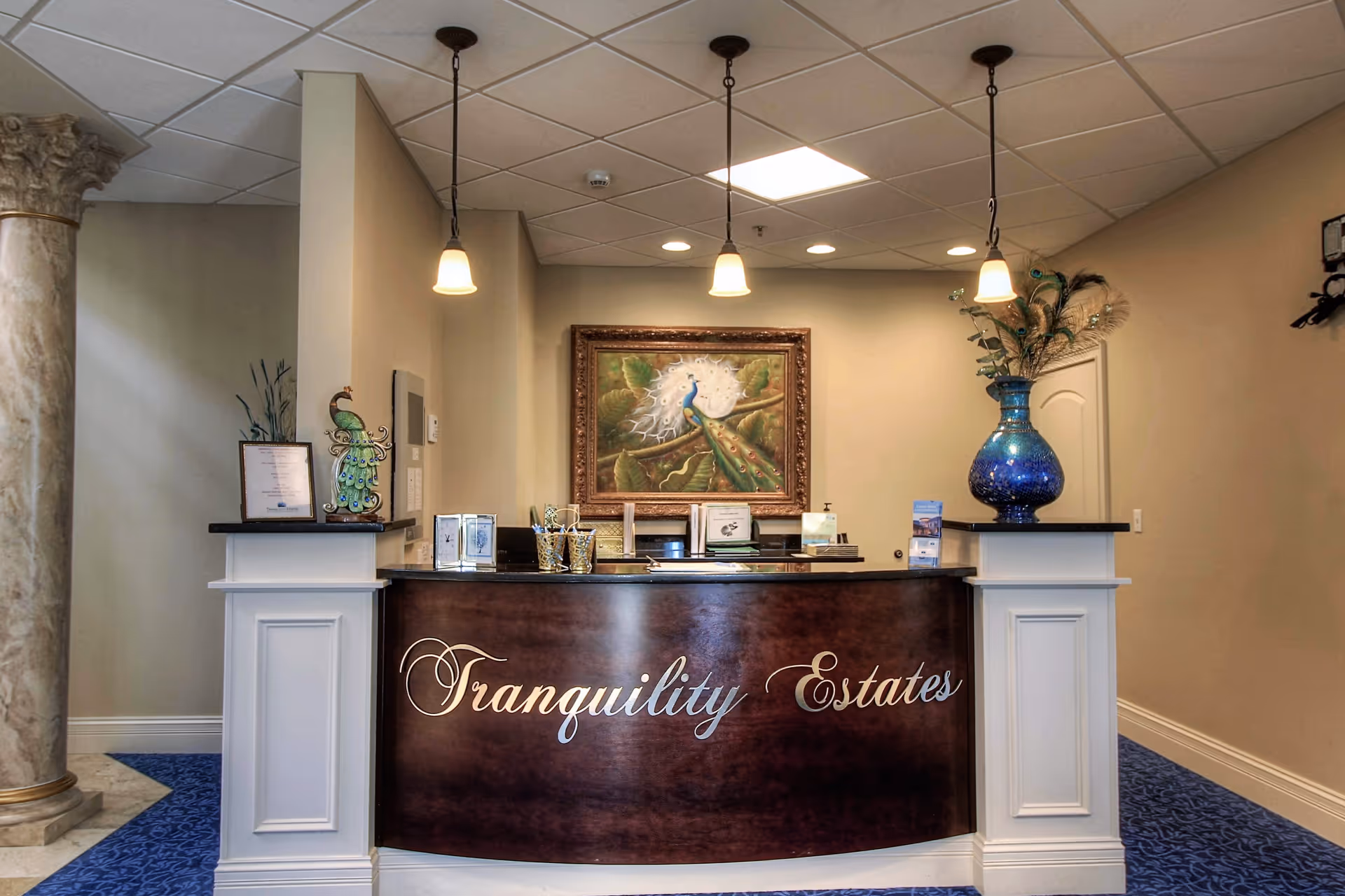 Reception desk area of Tranquility Estates with a dark wood front panel displaying the facility name in elegant script. The desk is flanked by white pillars, with a large blue vase holding decorative feathers on the right side and a peacock-themed decoration on the left. Above the desk hangs a framed painting of a peacock. The ceiling has recessed lighting and three pendant lights hanging above the desk. The floor is carpeted in blue with a patterned design.