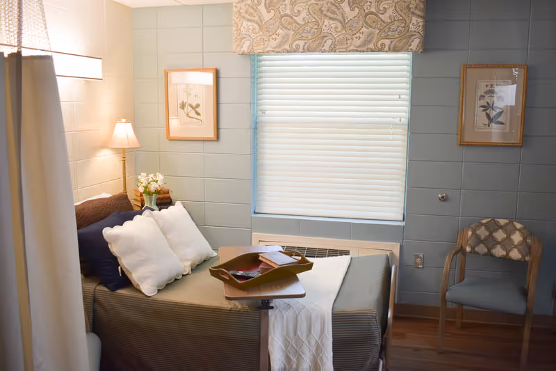 A small, cozy bedroom with a single bed covered in a brown bedspread, white pillows, and a white throw blanket. There is a wooden tray on the bed with a remote control and some papers. A floor lamp and a small vase with flowers are on a nightstand beside the bed. The walls are painted light blue with two framed botanical prints hanging on either side of a window with closed blinds and a patterned valance. A wooden chair with a patterned cushion is placed against the wall.