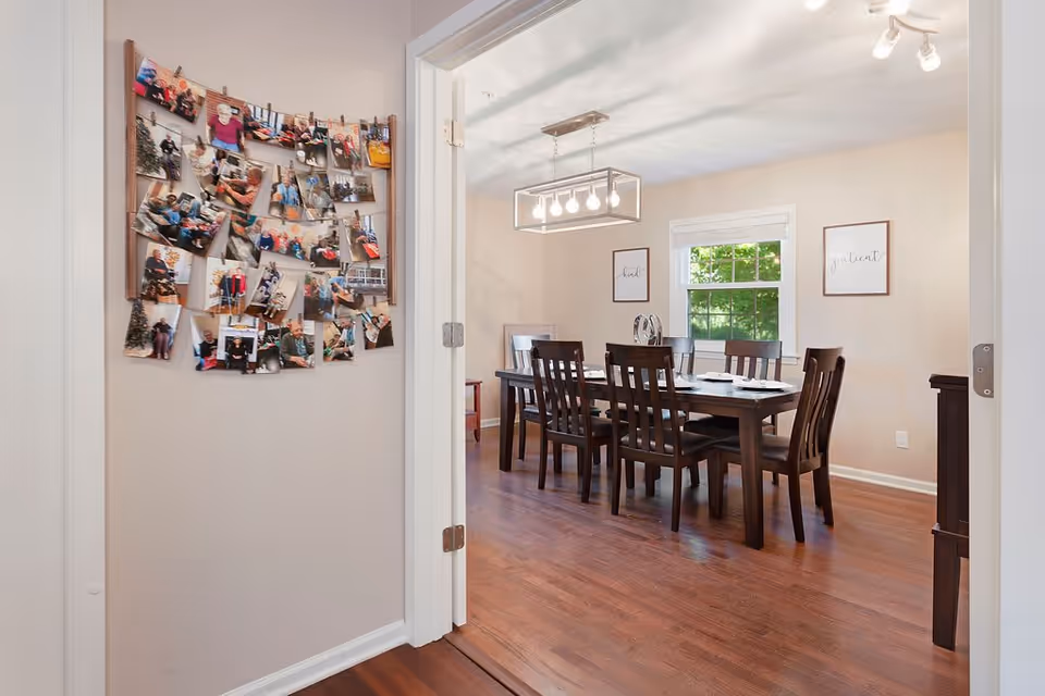 View through a doorway into a dining room with a dark wooden table and six matching chairs. The table is set with plates and napkins. On the wall to the left of the doorway, there is a display of numerous photos clipped to strings. The dining room has a window with white trim and two framed wall art pieces. The floor is wooden, and a modern rectangular light fixture hangs above the table.