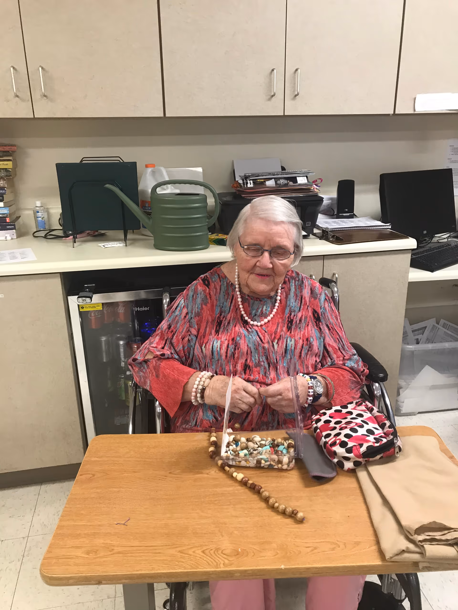 An elderly woman with white hair and glasses sits in a wheelchair at a wooden table. She is wearing a red and gray patterned top and pink pants, along with beaded bracelets and a necklace. On the table in front of her is a clear container with beads and a string of beads extending out. Behind her is a counter with various items including a green watering can, a computer monitor, and office supplies.