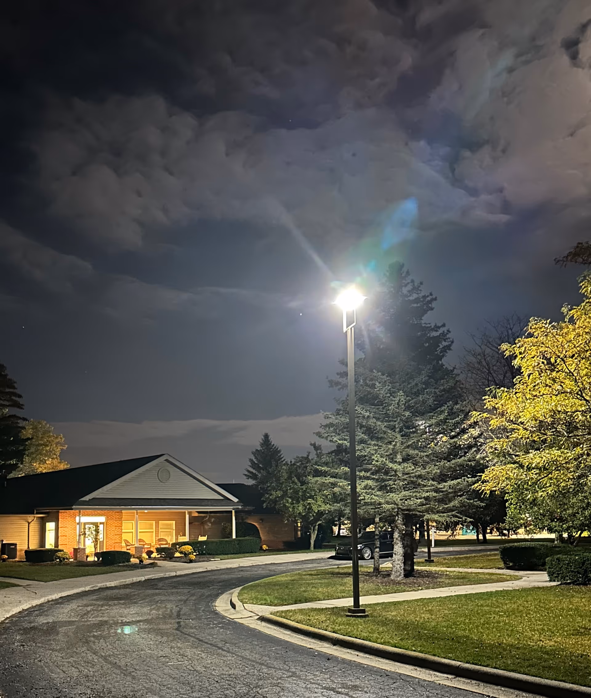Night view of a lit building entrance with a curved driveway, streetlamp, and trees under a cloudy sky.