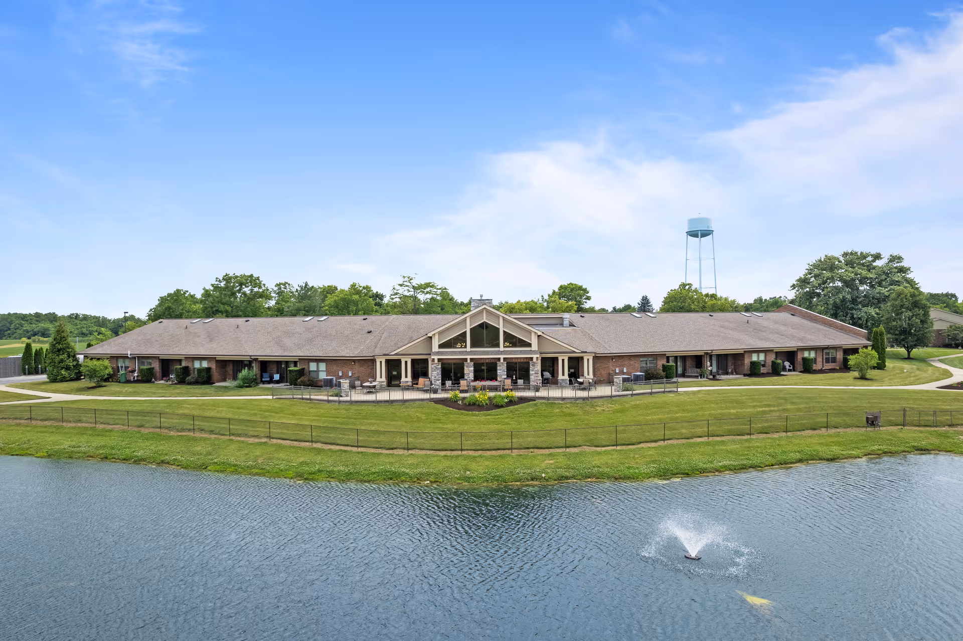 Single-story brick nursing facility building facing a pond with a fountain and a water tower in the background.