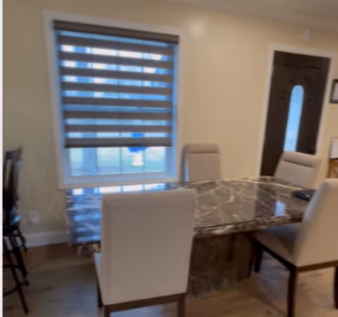 A dining area with a rectangular marble table surrounded by four beige upholstered chairs. There is a window with a striped roller blind partially down, and a dark-colored door with a frosted glass panel is visible in the background. The floor is wooden.