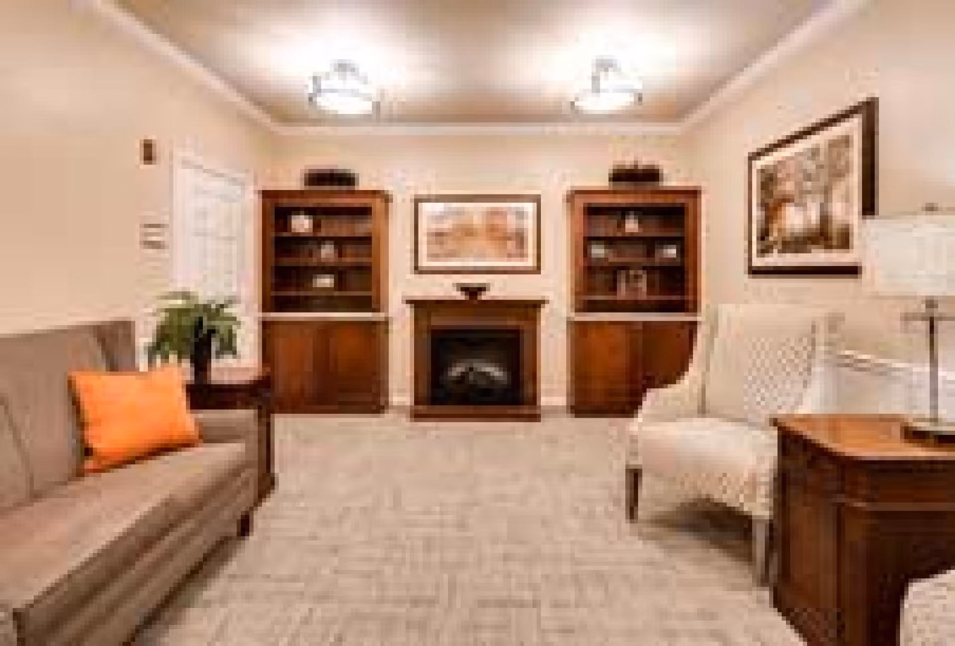 Cozy sitting room with a central fireplace between two wooden bookcases, a sofa with an orange pillow on the left and an armchair on the right.