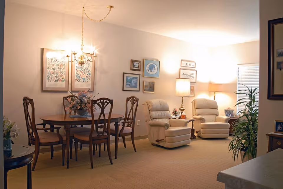 A cozy living and dining area featuring a wooden dining table with six chairs and a floral centerpiece, two beige recliner chairs with side tables and lamps, framed artwork on the walls, and a potted plant near the window with blinds.