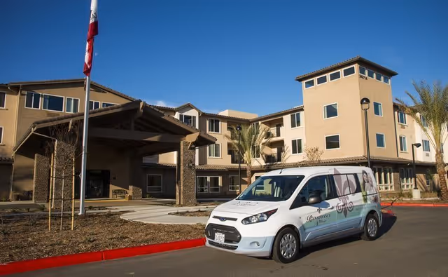 Exterior view of Renaissance Village Murrieta senior living facility with a white van parked in front. The building is multi-story with beige walls, several windows, and a covered entrance. There are palm trees and a clear blue sky in the background.